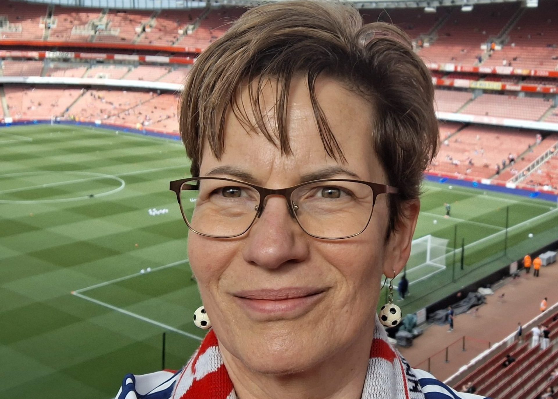 A person wearing glasses and a red-and-white scarf takes a selfie inside a large football stadium with red seats.